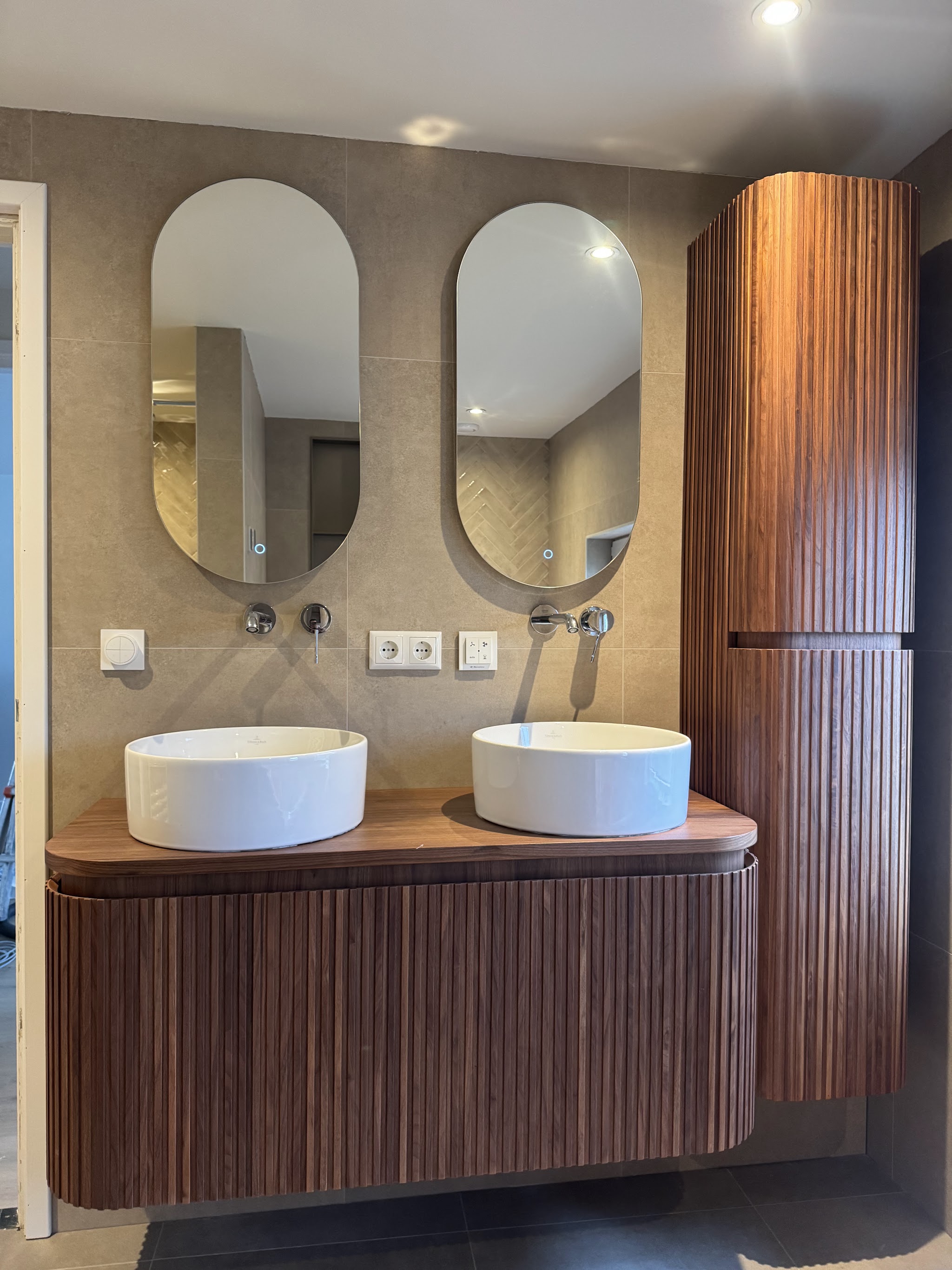 Badkamer met twee ovale spiegels, twee ronde waskommen op een houten badmeubel en een hoge kast